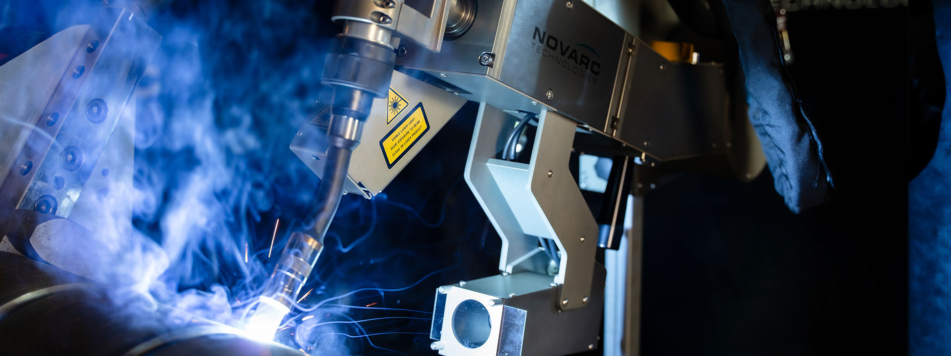 Close-up of a Novarc Spool Welding Robot welding a metal pipe, with blue arc light, smoke and sparks.